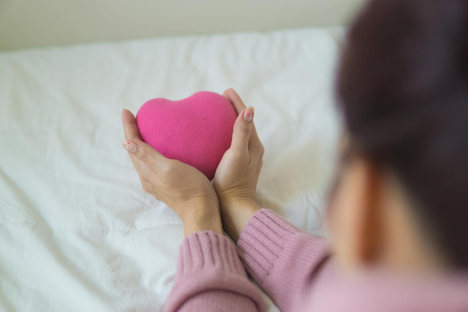 From above of unrecognizable female in sweater sitting on white blanket with pink heart shaped ex... (Photo by Puwadon Sang-ngern on Pexels)