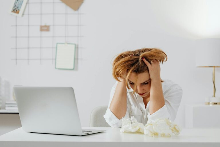 Frustrated woman in white shirt at desk with laptop, feeling overwhelmed. (Photo by www.kaboompics.com on Pexels)