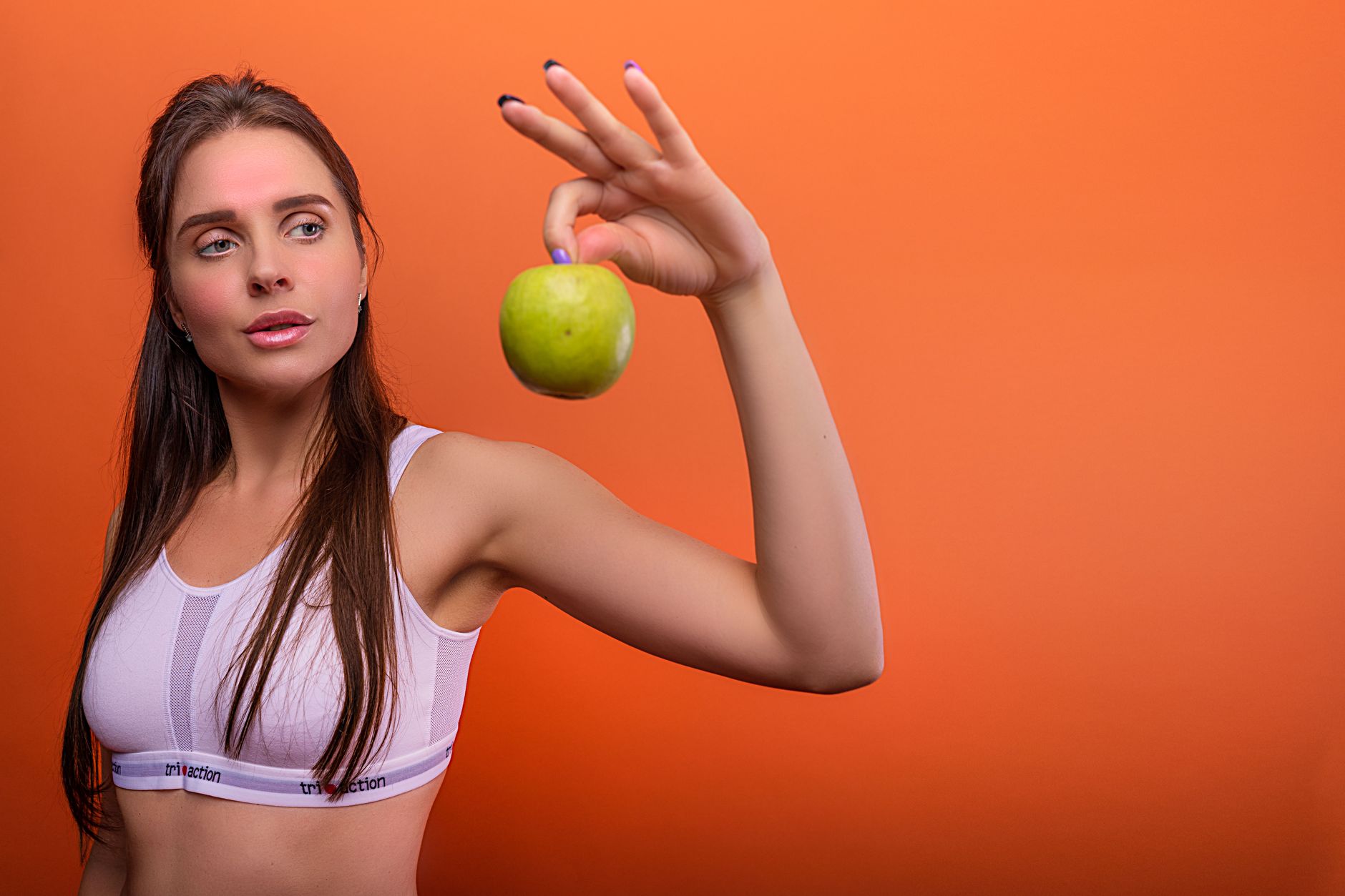 Young woman in activewear holding a green apple against orange background. (Photo by Atlantic Ambience on Pexels)