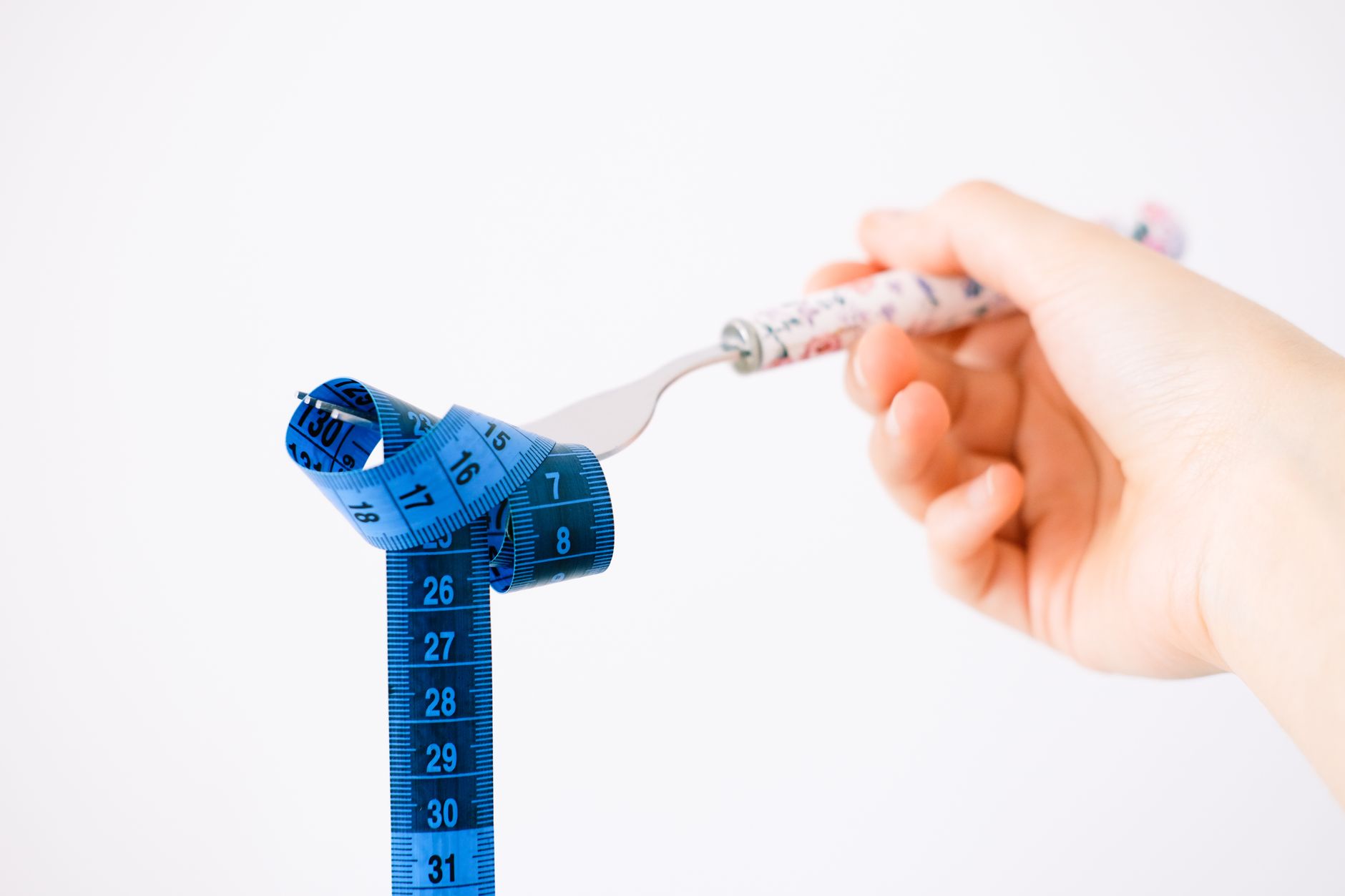 A hand holding a fork wrapped with a blue measuring tape suggesting diet or weight management. (Photo by Beyzaa Yurtkuran on Pexels)