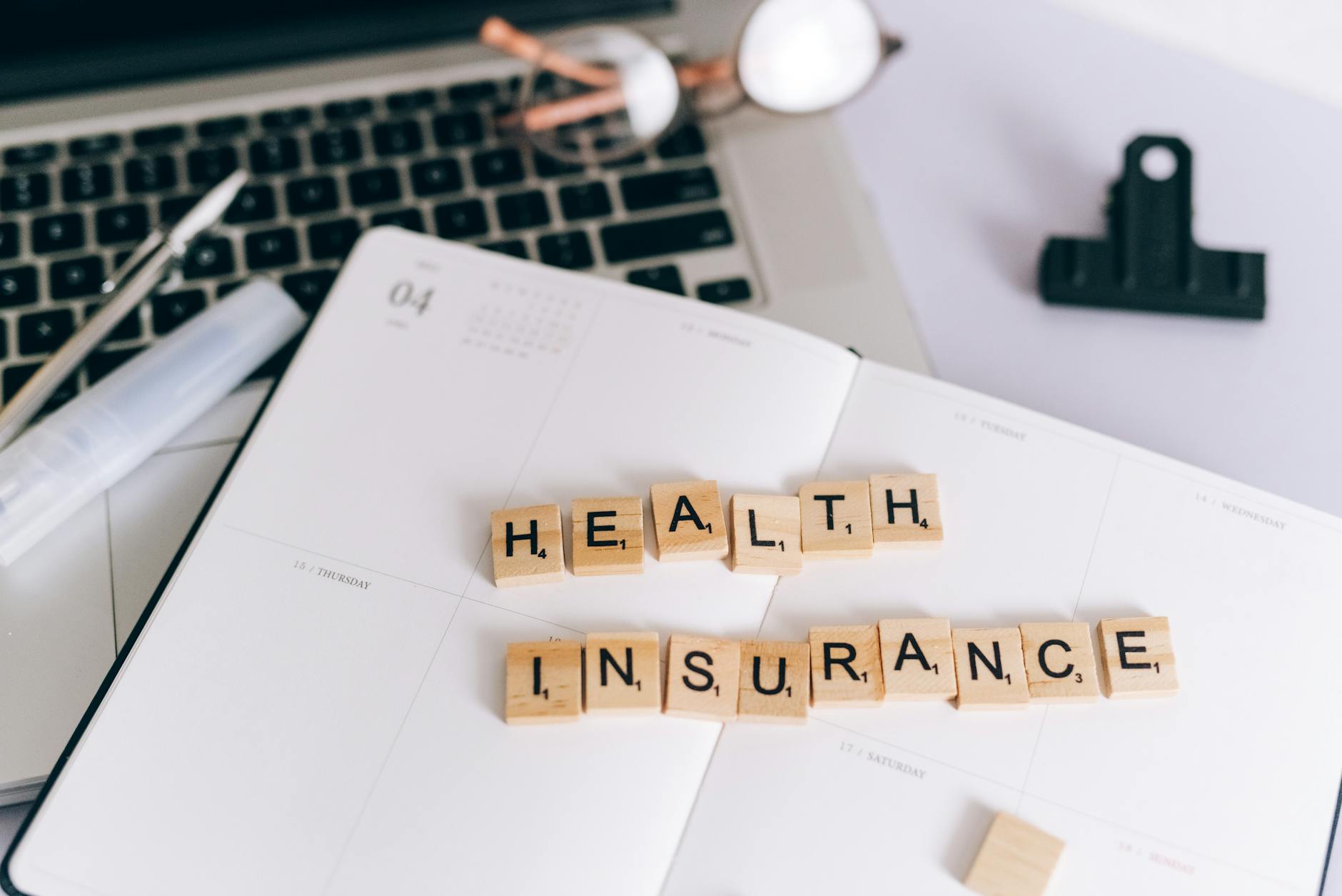 Close-up of Scrabble tiles spelling 'Health Insurance' on a planner with a laptop. (Photo by Leeloo The First on Pexels)